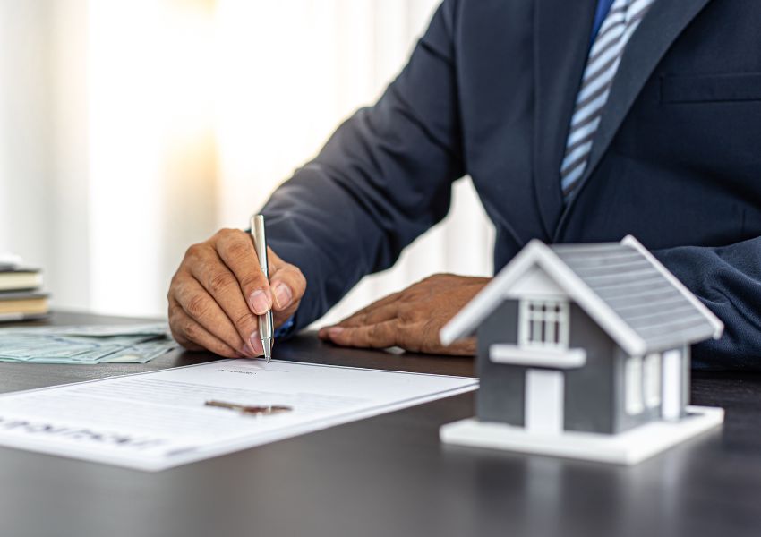 person in a suit writing on a piece of paper with a house figurine next to them